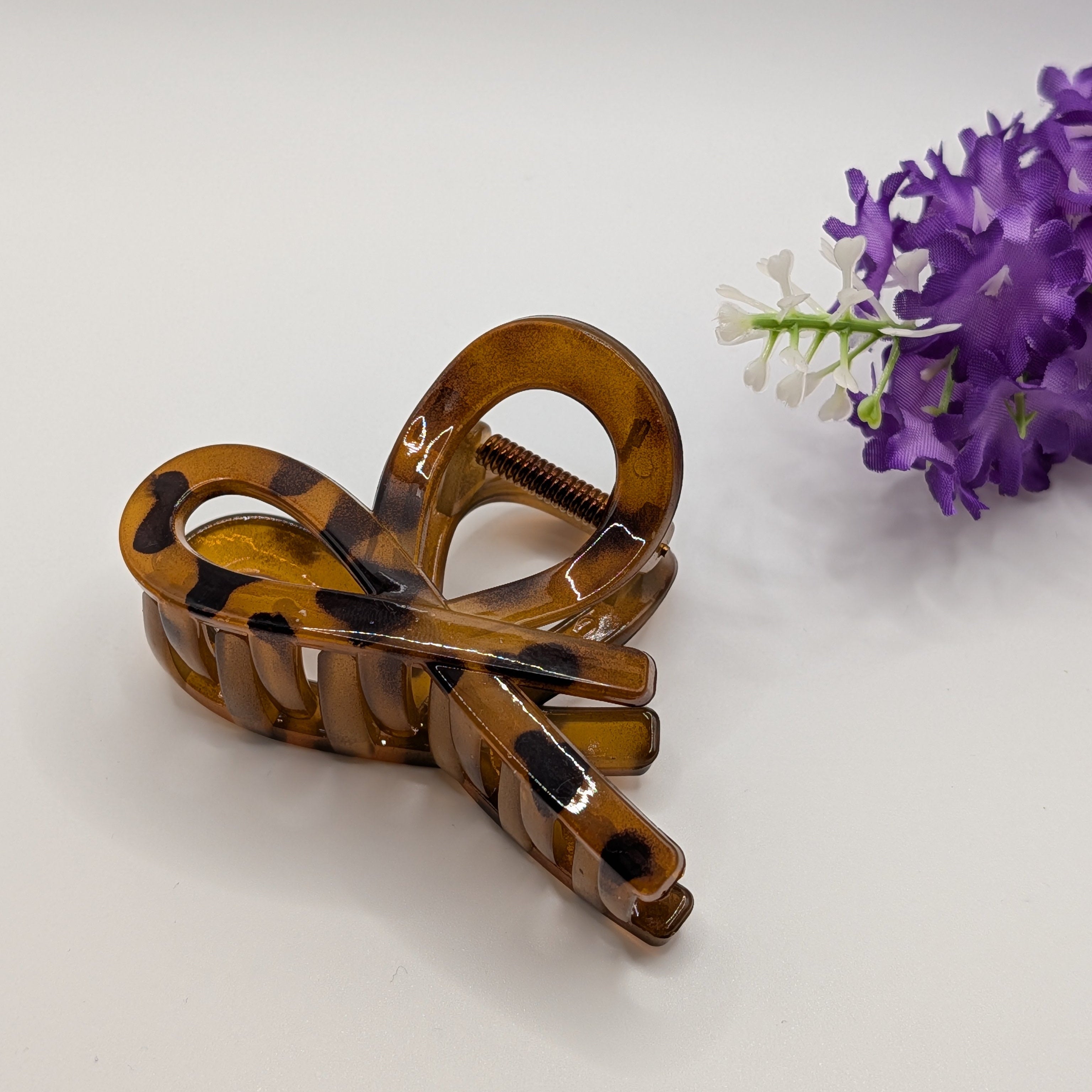 Braun - Elegant amber-colored bow hair clip placed next to a purple flower for lifestyle presentation.

Elegante bernsteinfarbene Haarschleifen-Klammer neben einer lila Blume zur Dekoration.