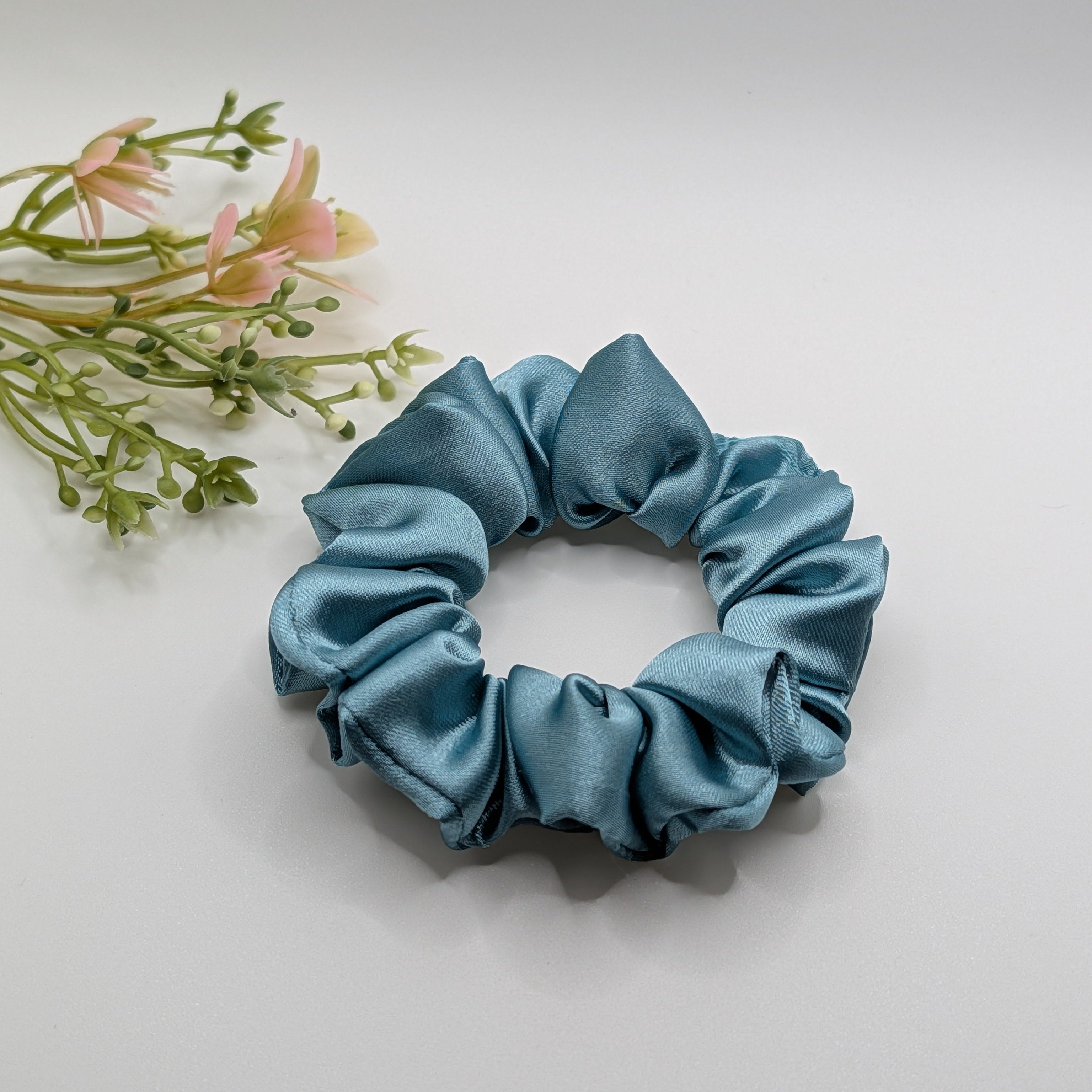 3er Set - Top view of a medium blue satin scrunchie on a white background.	

Draufsicht eines mittelgroßen blauen Satin-Haargummis auf weißem Hintergrund.