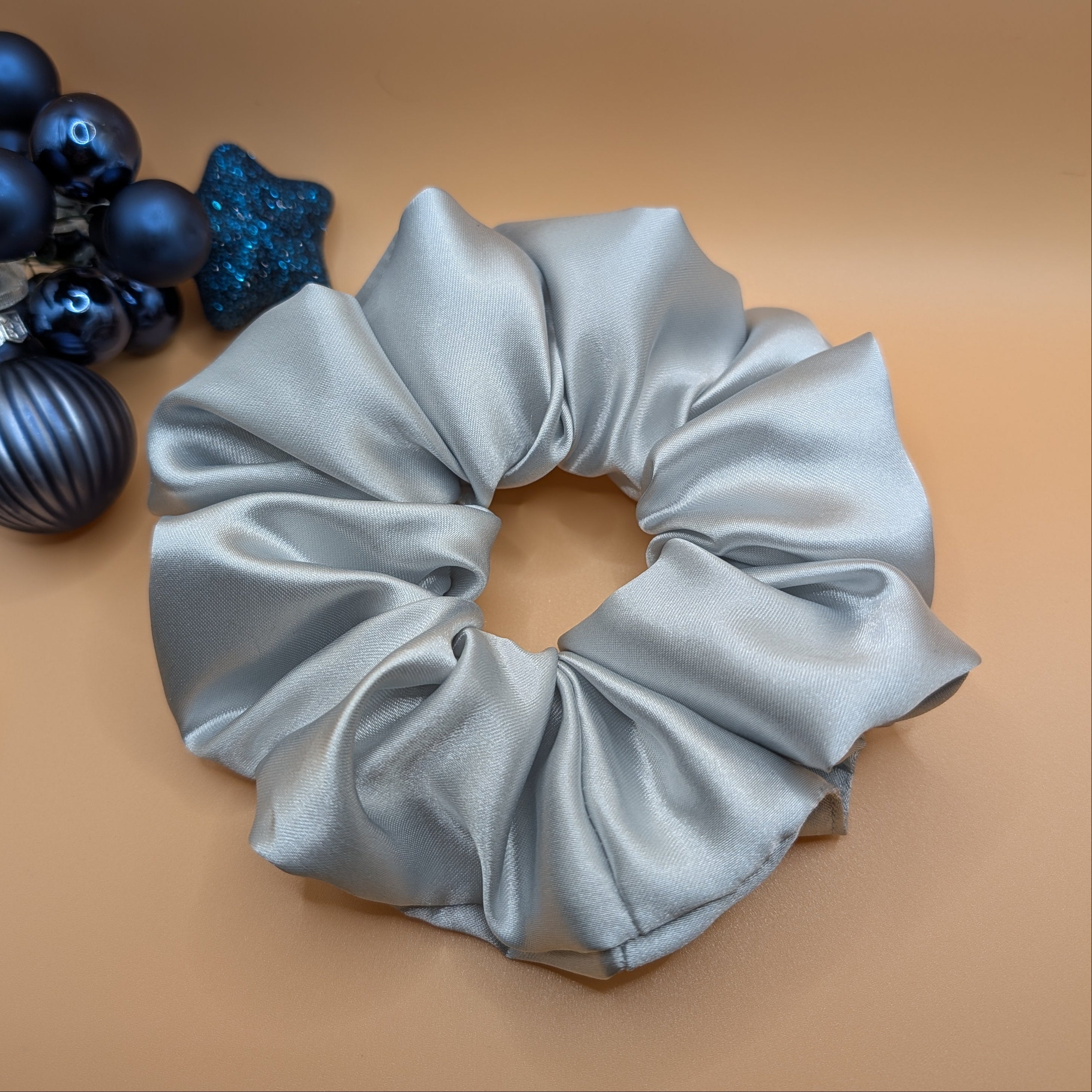 3er Set - Luxury silver satin scrunchie next to blue holiday ornaments.	

Luxuriöser silberner Satin-Scrunchie neben blauem Weihnachtsschmuck.
