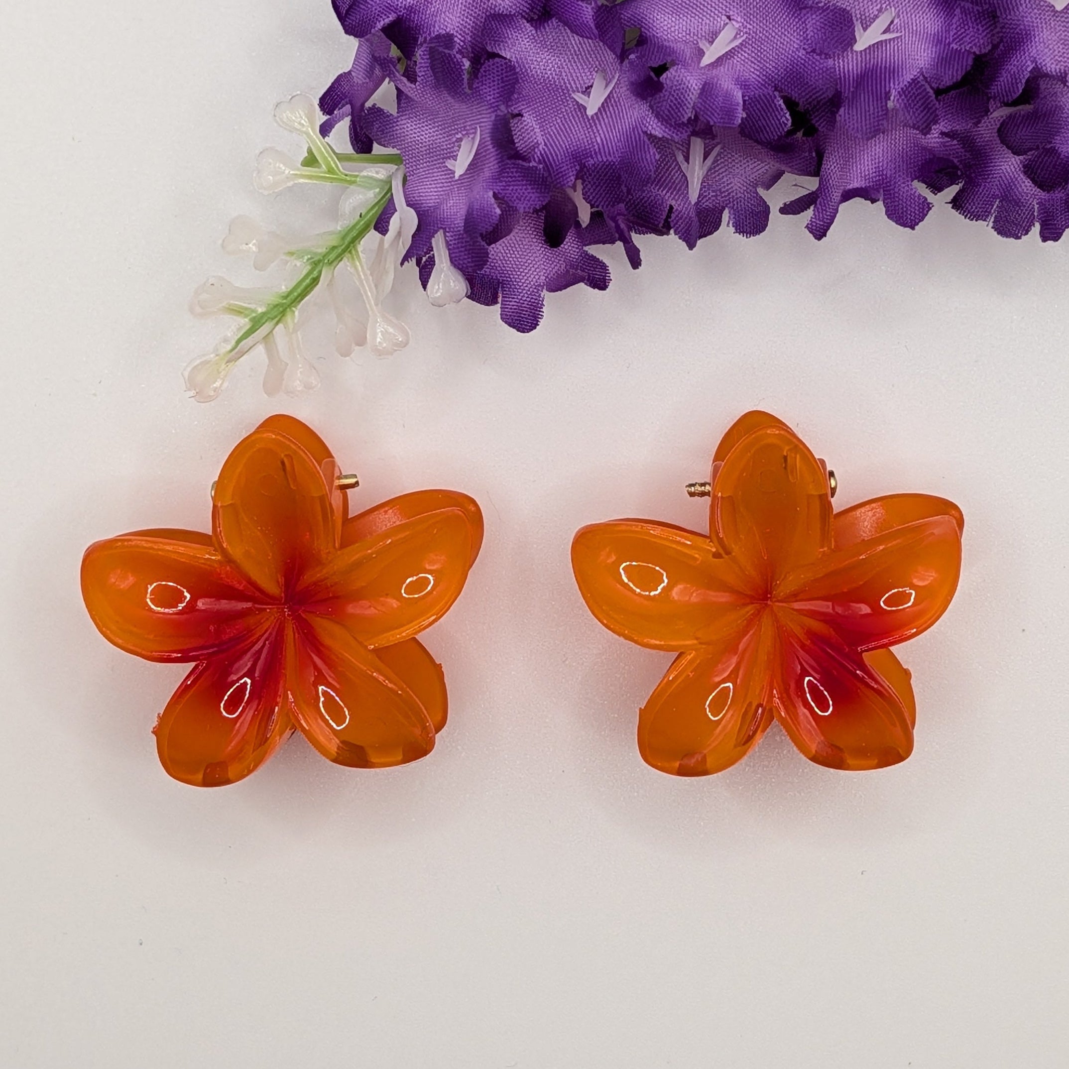 Leuchtendes Orange - Pair of small translucent orange flower hair clips on white background with purple flora.

Paar kleine, orangefarbene Blumen-Haarclips auf weißem Hintergrund mit lila Blumen.