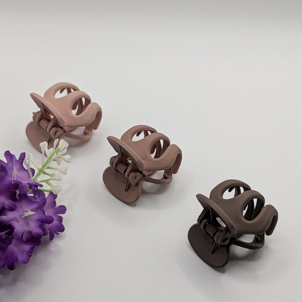 Three matte rounded hair clips arranged diagonally next to purple flowers on a white surface.	

Drei matte, abgerundete Haarklammern, diagonal angeordnet neben lila Blumen auf einer weißen Fläche.