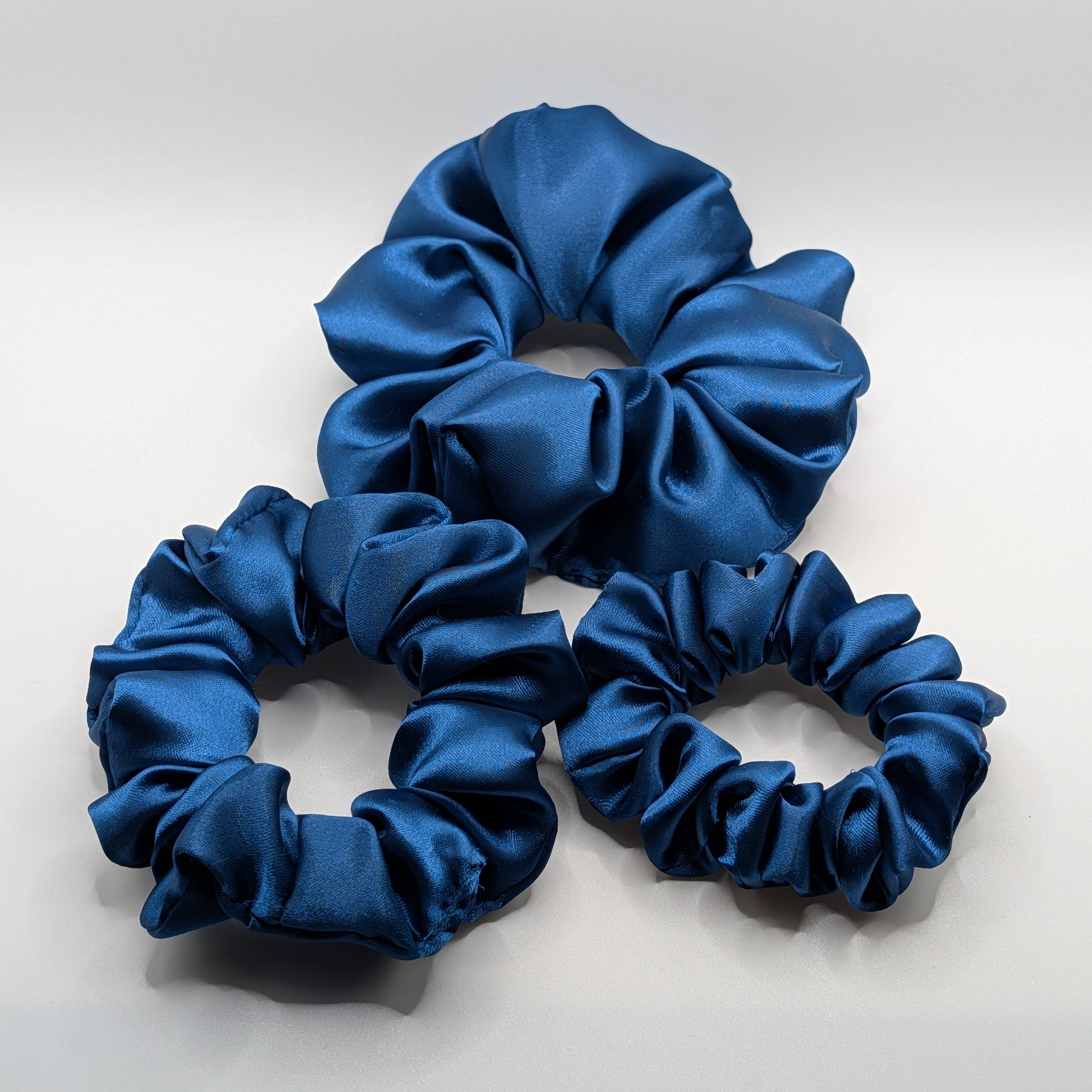3er Set - 3-piece navy blue satin scrunchie set showing large, medium, and small sizes.	

3-teiliges Set blaue Satin Haargummis in den Größen Groß, Mittel und Klein.