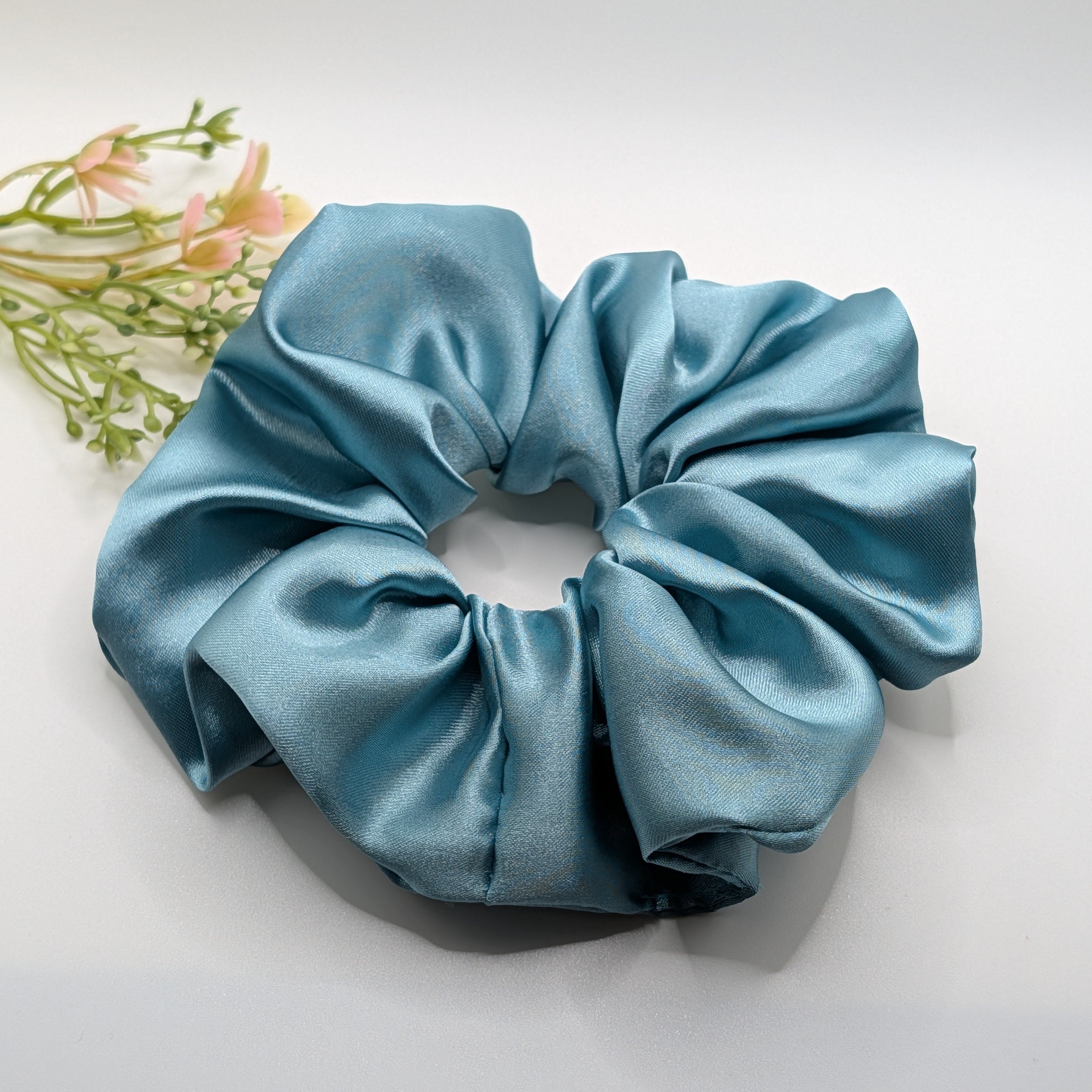 3er Set- Oversized blue satin scrunchie next to decorative pink flowers.	

Übergroßes blaues Satin-Haargummi neben dekorativen rosa Blumen.